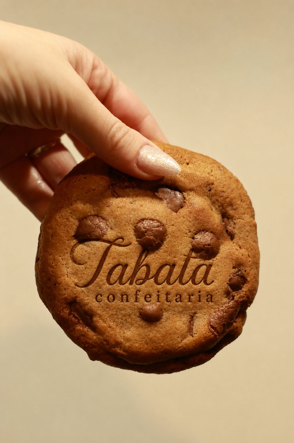 Logo Tabata Confeitaria sobre um cookie com gotas de chocolate, segurado pela mão